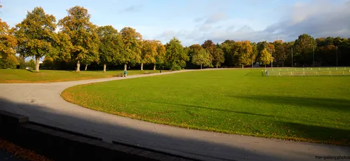 This landscape photograph captures Queen's Park in the United Kingdom during an autumn morning, showcasing the seasonal transformation of nature. The main subject of the image is Queen's Park, with well-maintained grassy areas and gently curving paths that wind through the park. The trees, displaying a mix of green and golden foliage, line the edges of the paths and reflect the colourful changes typical of autumn. Soft morning light casts long shadows and accentuates the vibrant colours of the park, highlighting both the trees and the open grassy space. Queen's Park is a notable green area in the United Kingdom, and the image effectively conveys its tranquil atmosphere and the beauty of its natural surroundings.